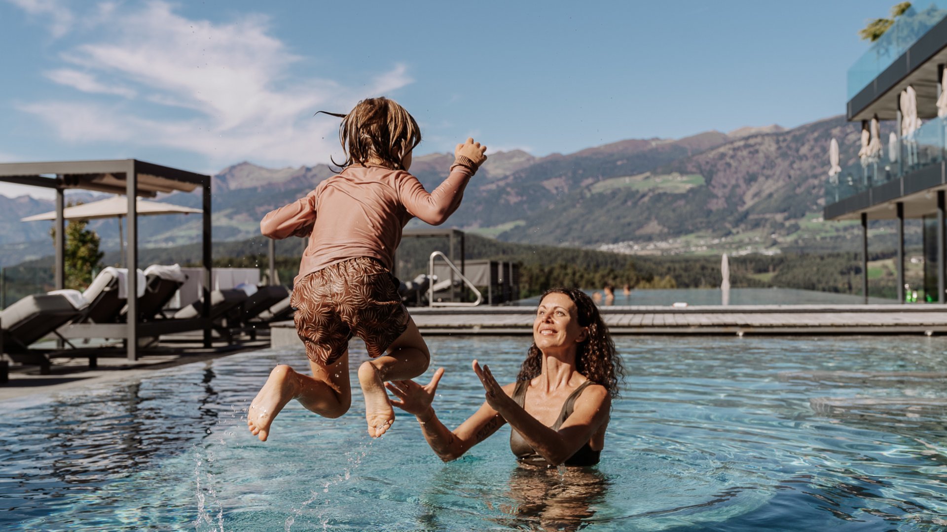 Kind springt in den Pool zu Frau, Berge im Hintergrund